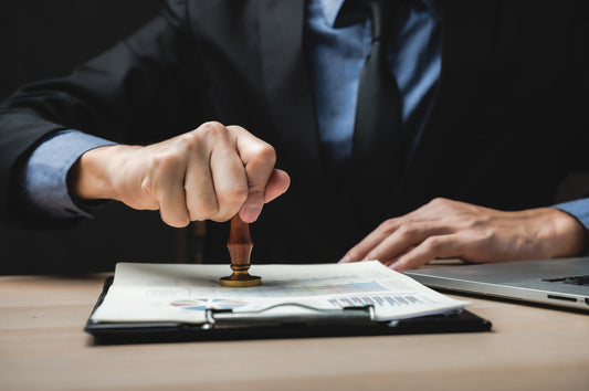 A businessperson in a suit stamps a document on a desk with charts and a laptop, symbolizing approval or authorization.