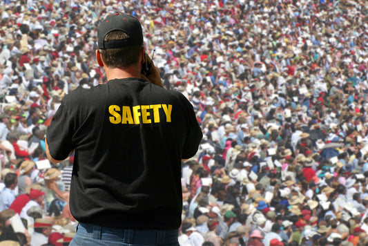 A safety officer wearing a black shirt monitors a large crowd of people gathered outdoors at a public event.
