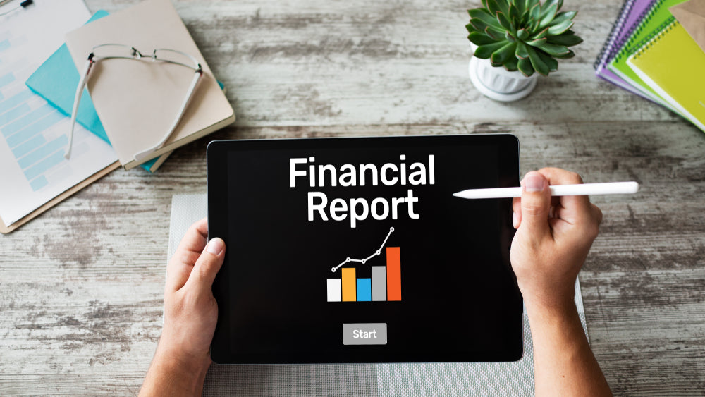 Person holding a tablet displaying 'Financial Report' with a bar and line chart, alongside a stylus, documents, and a plant.