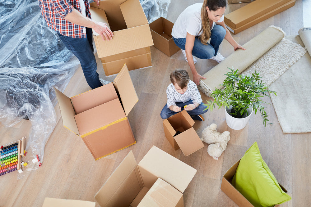 Family unpacking cardboard boxes in a new home, with a child playing, a plant, and household items scattered around.