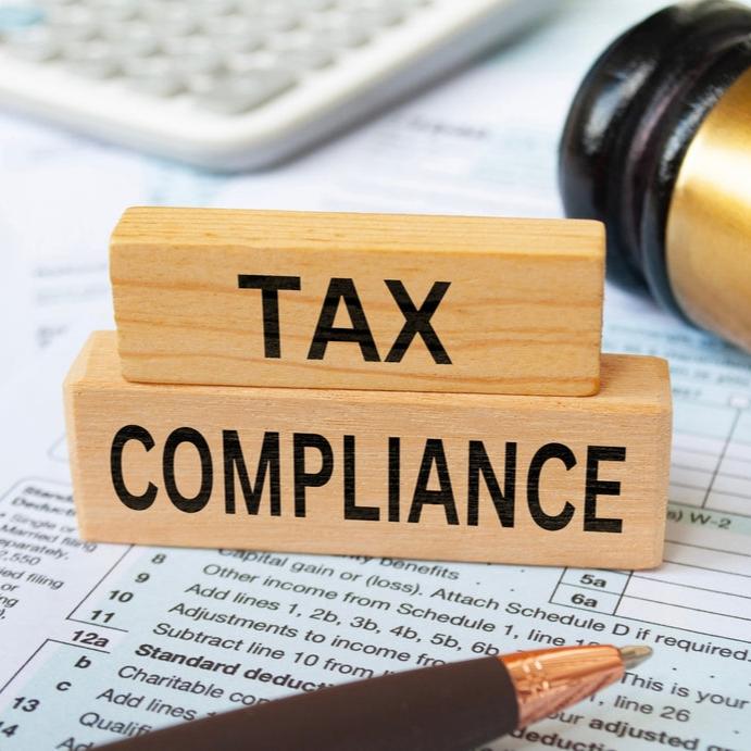 Wooden blocks reading 'Tax Compliance' placed on tax forms with a pen, gavel, and calculator in the background.