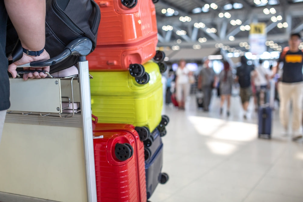 Traveler pushing a luggage cart loaded with colorful suitcases in a busy airport terminal with people in the background.