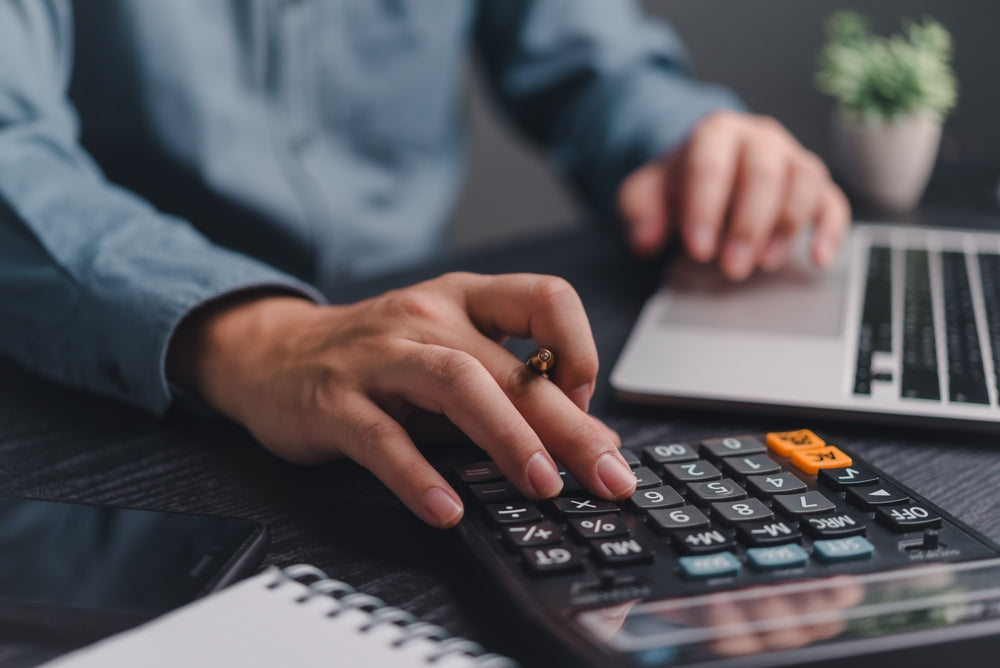 Close-up of a person using a calculator and laptop on a desk, with a notepad, phone, and small plant in the background.