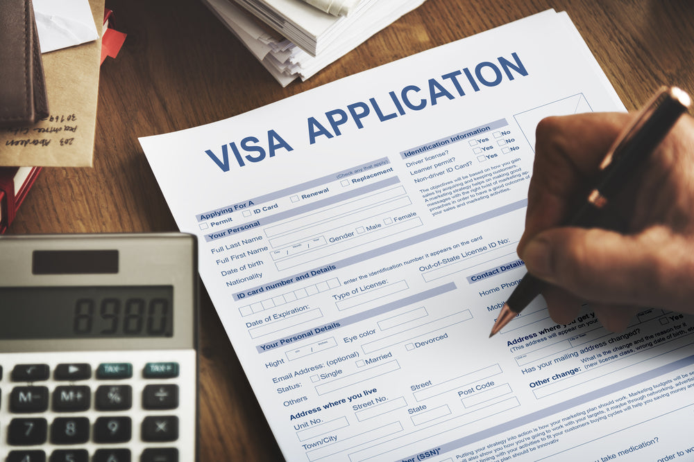 Close-up of a visa application form being filled out by hand, with a calculator and documents on a wooden desk.